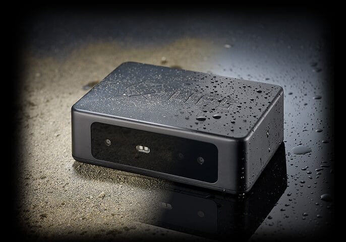 A compact black electronic device with a water-resistant design, featuring water droplets on its surface, placed on a wet reflective surface.