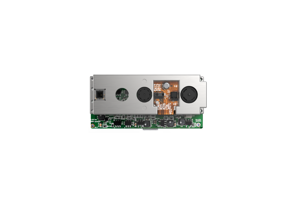 A compact electronic module featuring a metal casing with multiple circular and rectangular openings, exposing internal components and sensors. The lower section reveals a green circuit board with intricate wiring and small electronic parts.