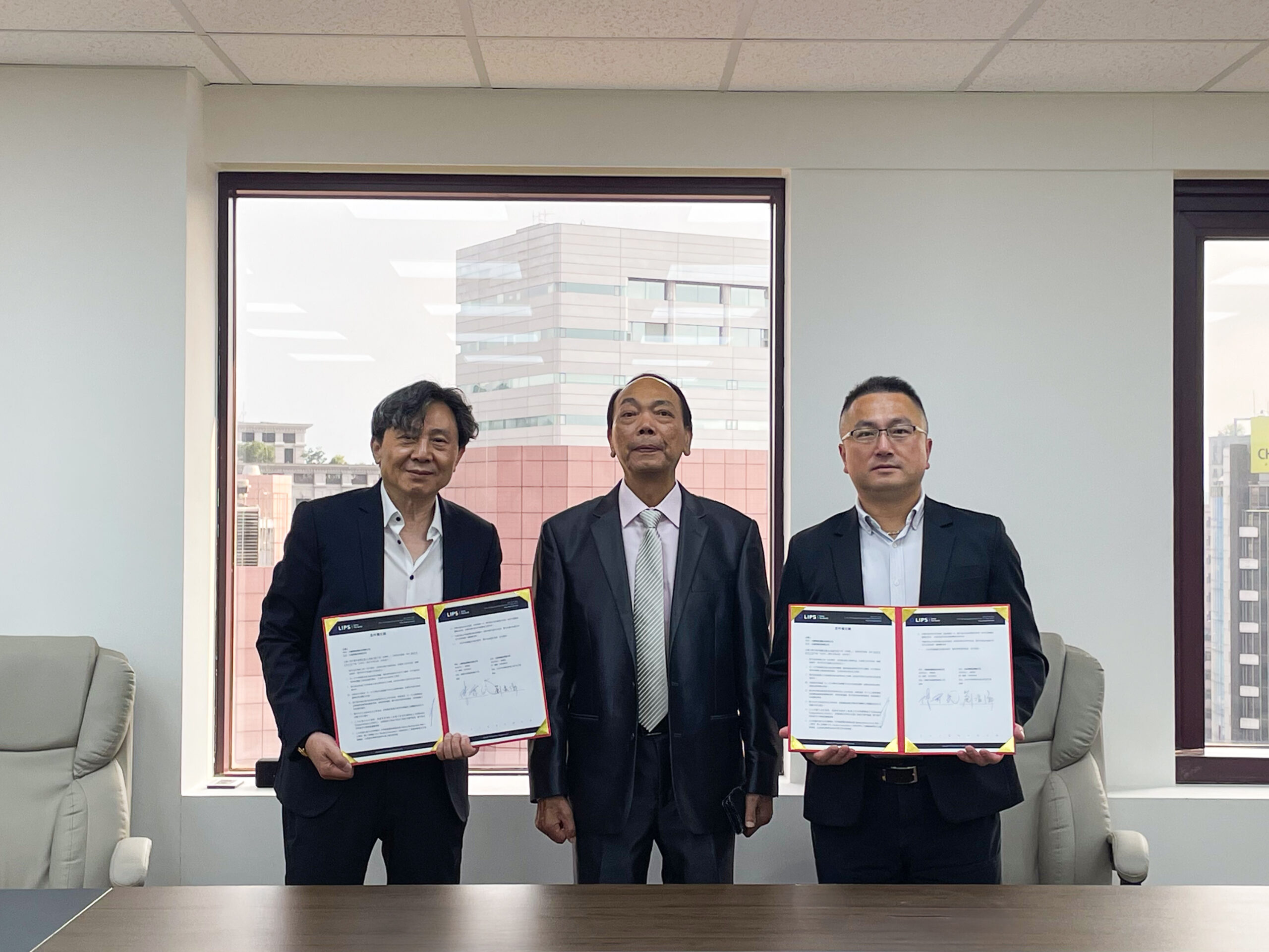 Three executives in business suits standing in an office and holding signed partnership documents in open folders during a formal ceremony, with a city building visible through the window behind them.